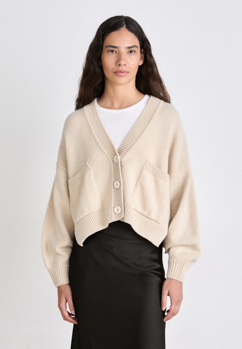 Beige cropped cardigan with V-neckline, ribbed edges, and two front pockets. Features three large buttons and soft knit texture.
