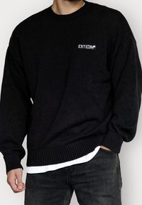 Pull en maille noire avec poignets et ourlet côtelés, présentant un petit logo blanc "ESTEEM" sur le côté gauche de la poitrine. Porté par-dessus une chemise blanche.