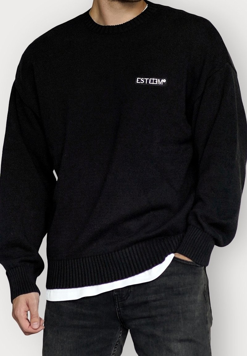 Pull en maille noire avec poignets et ourlet côtelés, présentant un petit logo blanc "ESTEEM" sur le côté gauche de la poitrine. Porté par-dessus une chemise blanche.