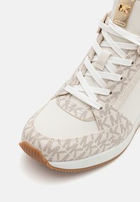 Sneaker atletica beige con tomaia in tessuto a fantasia, lacci bianchi e suola in gomma. Presenta dettagli con logo e una finitura texturizzata.
