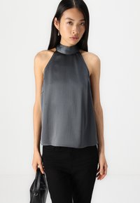 Haut halter gris en tissu lisse et texturé, avec un col haut et une coupe ample, associé à un pantalon noir et tenant un petit sac noir.
