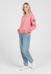 Felpa rosa con grafiche sulle maniche, abbinata a jeans azzurri chiari e sneakers bianche. Tessuto morbido, vestibilità rilassata, stile casual.