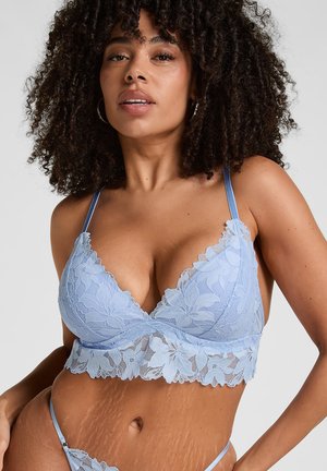 Frau mit lockigem Haar, die ein hellblaues, florales Spitzen-Bralette und passende Unterwäsche vor einem einfarbigen Hintergrund trägt.