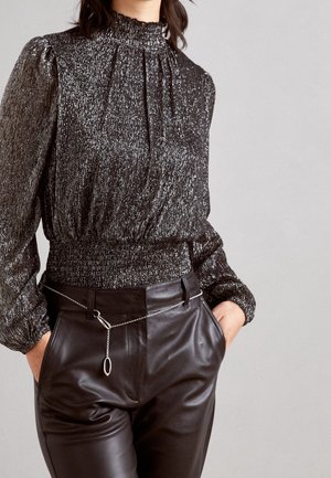 Femme portant un chemisier noir brillant à manches longues avec col montant et pantalon en cuir noir avec ceinture chaîne argentée, mains dans les poches.