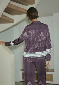Lilla bluse med blomsterprint, hvide accentstriber og tekstdetaljer. Glat stof, afslappet pasform, synlige folder bagpå, kombineret med matchende bukser.