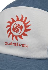 Gorro azul y blanco hecho de tejido texturizado, con un gráfico de un sol rojo y un logotipo con la palabra "Quiksilver" debajo.