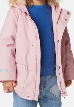 Dziecko w rozpiętej jasnoróżowej kurtce z kapturem, pod którą ma niebieski sweter i jeansy z łatą w kształcie serca na lewej udzie.