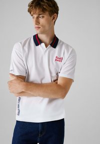 Hvid polo skjorte med marineblå og rød stribet krave, med "Red Bull RACING" logo på brystet og "GIVES YOU WINGS" tekst på siden af kanten.