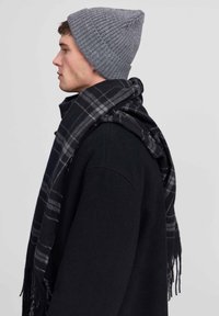 Szara, prążkowana czapka beanie w połączeniu z czarno-szarym, kratkowanym szalem z postrzępionymi brzegami. Model widziany z boku, noszący czarny płaszcz.