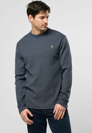 Pull en maille texturée bleu marine à manches longues avec un col rond et un petit logo doré sur la poitrine. Porté avec un jean foncé.
