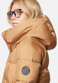 Beżowa kurtka puchowa z wysokim kołnierzem, cechująca się błyszczącym wykończeniem, pikowaną teksturą oraz okrągłą łatą z logo na lewym rękawie.