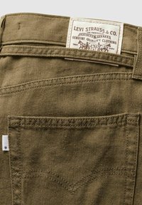 Poche arrière d'un jean Levi's marron avec une étiquette blanche Levi Strauss & Co. cousue au-dessus de la poche et l'onglet rouge Levi's partiellement visible à gauche.
