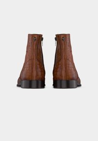 Ζευγάρι από καφέ δερμάτινα μποτάκια αστραγάλου με υφή κροκοδείλου, με μαύρα φερμουάρ σε σχήμα κρανίου, που φαίνονται από πίσω πάνω σε λευκό φόντο.