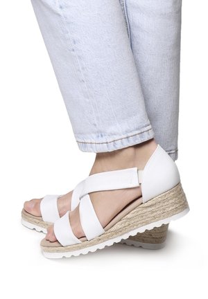 White wedge sandals with crossed straps, jute-wrapped midsole, and a textured rubber sole, paired with light blue jeans.