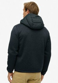 Giacca in pile grigio scuro con cappuccio quilted verde scuro, caratterizzata da una vestibilità morbida e un tessuto testurizzato. Il design include un orlo dritto e polsini elasticizzati.