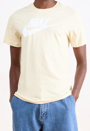 Mann trägt hellbeiges Nike T-Shirt mit weißem Logo und blauer Jeans, Hände in den Taschen, Vorderansicht, beschnitten am Hals und an den Oberschenkeln.