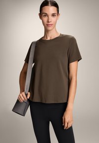 Olivnozeleni bombažni t-shirt s kratkimi rokavi in okroglim ovratnikom; v kombinaciji z črnimi pajkicami. Model drži sivo torbo z naramnico.