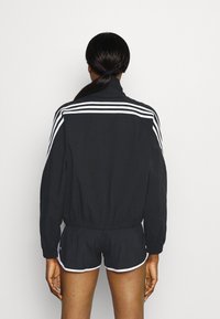 Schwarze Sportjacke mit weißen Streifen an den Schultern, kombiniert mit schwarzen Shorts mit weißem Saum. Glattes Material, lässiger Schnitt.