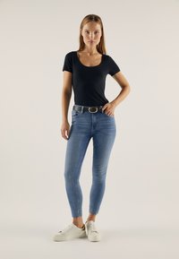 Černé upnuté tričko, modré skinny džíny s předními kapsami a bílé tenisky se zlatými akcenty, stojící na neutrálním pozadí.