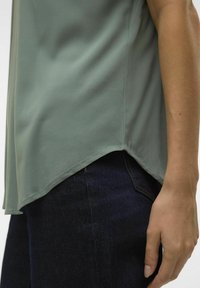 Top de jersey verde claro con dobladillo curvado, textura suave y mangas cortas, combinado con jeans de mezclilla oscura.