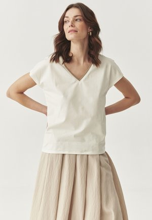 Top bianco a maniche corte con scollo a V, realizzato in tessuto morbido. Abbinato a una gonna beige fluida e plissettata. Look semplice e moderno.