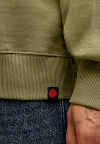 Oliwkowa bluza z ribbowanymi mankietami i dołem. Posiada czarną metkę z czerwonym okrągłym akcentem. Noszona na ciemnych dżinsach.