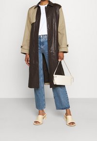 Kobieta w dwukolorowym długim płaszczu, białej bluzce, niebieskich dżinsach, beżowych sandałach na obcasie i trzymająca małą białą torebkę na jednolitym tle.