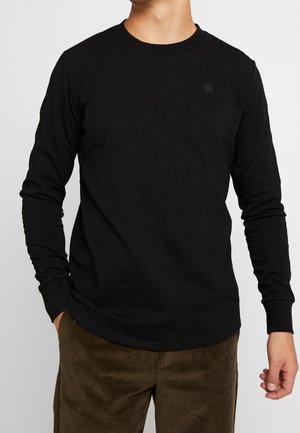 Homme portant une chemise noire à manches longues unie et un pantalon en velours côtelé marron, debout avec une main dans la poche sur un fond blanc.