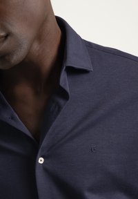 Camisa azul marino abotonada con textura suave, con cuello y un logo sutil bordado en el pecho. Detalle de botones blancos visible.