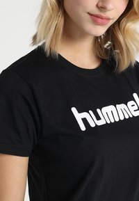 Blonde Frau trägt ein schwarzes T-Shirt mit weißem "hummel"-Logo auf der Brust, dargestellt von den Schultern bis zu den Lippen.