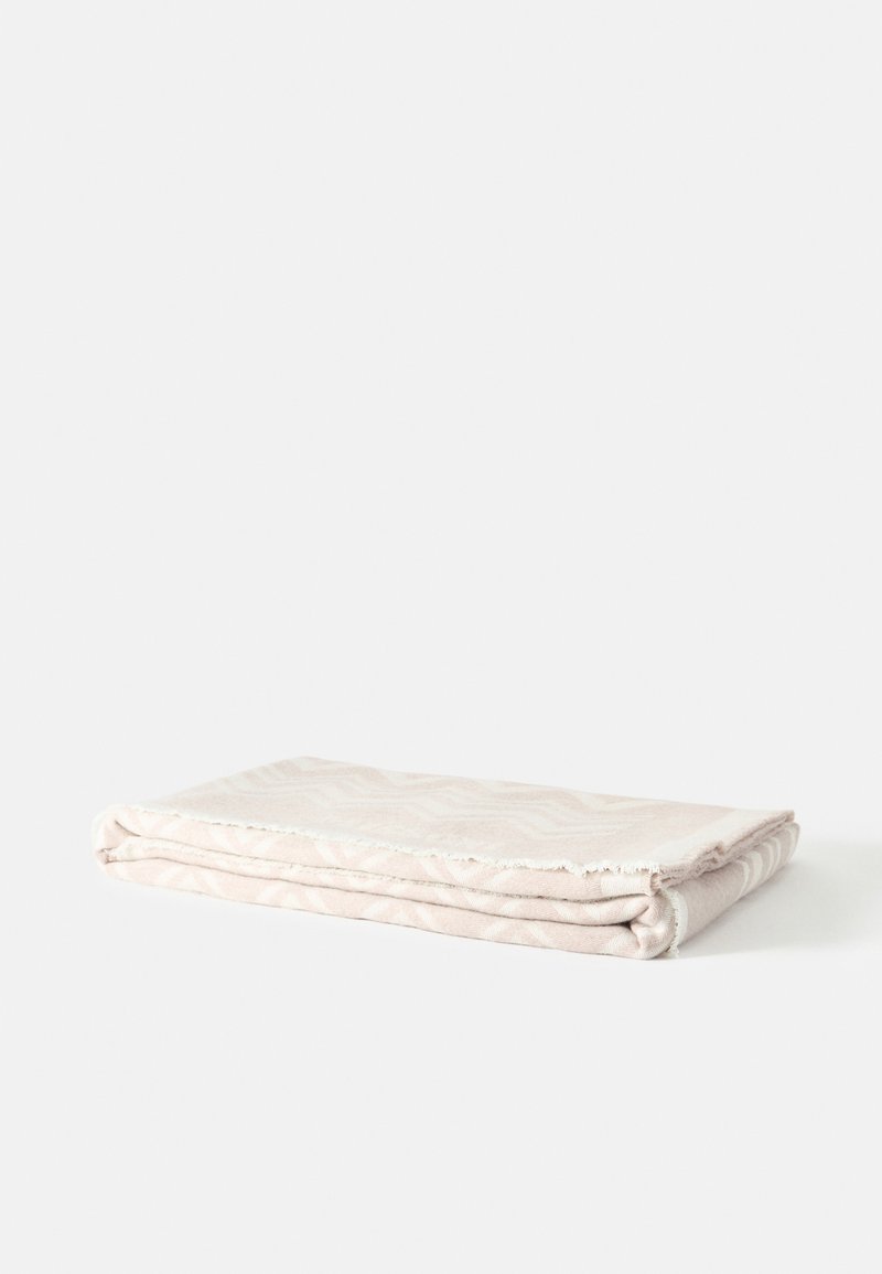 Couverture beige clair pliée avec des motifs subtils, posée à plat sur une surface blanche.
