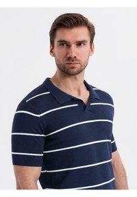 Námořnická modrá pletená polo tričko s bílými horizontálními pruhy, krátkými rukávy a klasickým límečkem. Obsahuje jemné logo na hrudi.