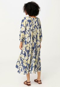 Pitkä kukkakuosisempi mekko, jossa on sinisiä ja keltaisia sävyjä, kokoontunut selkä, pitkät hihat, kerroksellinen helma ja kevyttä kangasta. Sandalet jalassa.