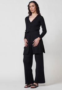 Cárdigan negro con canalé, de cuello en V, botones y un cinturón a juego, combinado con pantalones anchos de canalé. Sandalias con tiras negras.