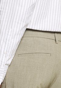 Lichtbeige broek met een gladde textuur en een enkele achterzak. Gecombineerd met een witte shirt met fijne grijze strepen.