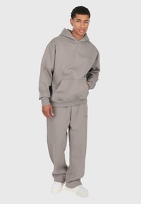 Junger Mann in einem lockeren grauen Kapuzenpullover und passenden Jogginghosen mit weißen Sneakern, der mit einer Hand in der Tasche vor einem schlichten Hintergrund steht.