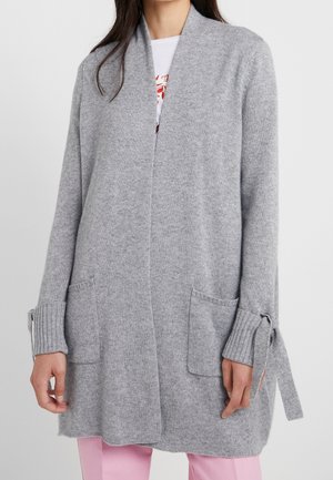 Frau trägt eine hellgraue, offene Strickjacke mit Taschen und gebundenen Bündchen über einem weißen Hemd und rosa Hose.