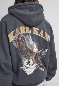 Felpa grigia con grafica di un'aquila, testo "KARL KANI" e un banner con "FLY". Realizzata in materiale morbido con una vestibilità ampia e rilassata.