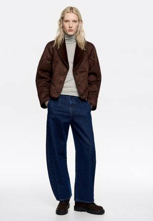 Mujer con cabello largo rubio que lleva una chaqueta marrón oversized, un jersey de cuello alto gris, vaqueros anchos azul oscuro y zapatos marrón oscuro, posando sobre un fondo blanco.