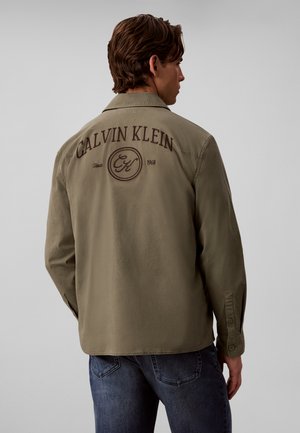 Mand iført olivengrøn Calvin Klein jakke med logo og tekst på bagsiden, kombineret med blå jeans, vendt væk mod en ensfarvet baggrund.