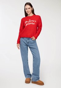 Czerwony sweter z wełny z białym napisem "Na dobrej liście Świętego Mikołaja", zestawiony z jasnoniebieskimi dżinsami i brązowymi botkami za kostkę.