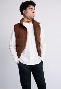 Brah vestska z zadrgo in stranskimi žepi, oblečena čez belo srajco. Črne hlače dopolnjujejo outfit.