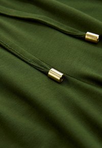 Grøn bomuldsstof med en glat tekstur, der har metallic guld hardware detaljer og snore til justerbar styling.