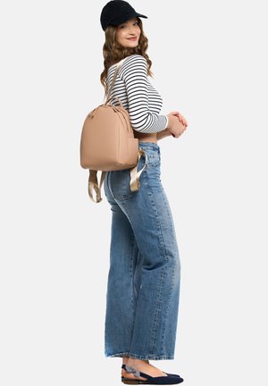 Beige backpack with textured material, rounded shape, adjustable gold-toned straps, and side pocket. Model wears a striped top and blue jeans.