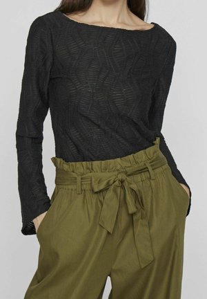 Femme portant un haut noir texturé à manches longues et un pantalon taille haute vert olive avec une ceinture en tissu nouée et des poches.