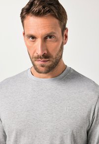 Pilkas medvilnės megztinis su apvalia kaklo iškirpte, turintis subtilų logotipą ant krūtinės ir glotnią tekstūrą. Artimas vyriško modelio viršutinės kūno dalies kadras.