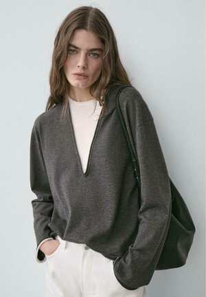 Donna con capelli castani lunghi che indossa un maglione grigio scuro sopra una camicia bianca e pantaloni bianchi, porta una borsa nera a tracolla, davanti a un muro semplice.