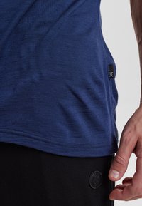Maglietta blu navy a maniche corte in tessuto leggero, con una sottile trama e etichetta laterale. Indossata sopra pantaloni neri con un'etichetta rotonda.