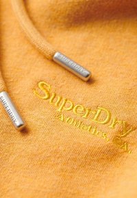Oranžová mikina s texturovaným povrchem; obsahuje žlutě vyšívané logo "SuperDry" a stříbrné kovové koncovky na tkaničkách.