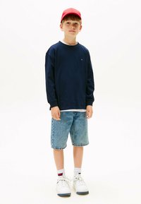 Marineblaues Langarmshirt über einem weißen T-Shirt, hellblaue Denim-Shorts, rotes Cap und weiße Sneaker mit grauen Akzenten.